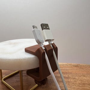 May include: A brown, C-shaped plastic cable organizer clip attached to a circular white stone surface. The clip features two vertical slots holding two white charging cables, one with a USB-C connector labeled onn. and one with a standard USB-A connector.