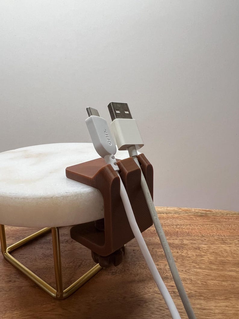 May include: A brown, C-shaped plastic cable organizer clip attached to a circular white stone surface. The clip features two vertical slots holding two white charging cables, one with a USB-C connector labeled onn. and one with a standard USB-A connector.