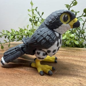 May include: A detailed, 3D-printed figurine of a falcon. The bird has gray and white feathers, yellow feet, and a yellow beak. The eyes are large and cartoonish. The figurine is posed on a wooden surface with green foliage in the background.