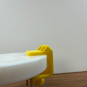 May include: A bright yellow C-clamp made of plastic, featuring a top flat surface and a threaded tightening knob at the bottom. The clamp is securely attached to the edge of a round, white marble tabletop that is supported by a thin gold metal frame.