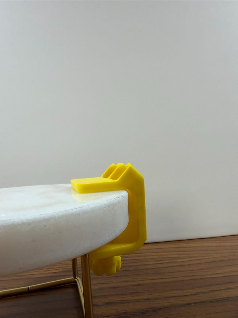 May include: A bright yellow C-clamp made of plastic, featuring a top flat surface and a threaded tightening knob at the bottom. The clamp is securely attached to the edge of a round, white marble tabletop that is supported by a thin gold metal frame.