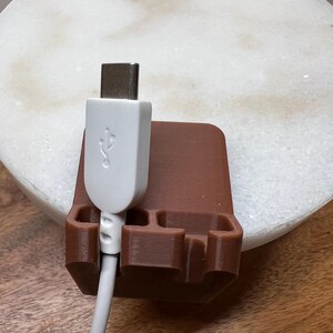 May include: A brown, 3D-printed cable organizer stand featuring three distinct slots for holding cords. A white USB-C cable is inserted into the leftmost slot, with the connector head facing upward, showcasing the standard USB symbol on its casing.