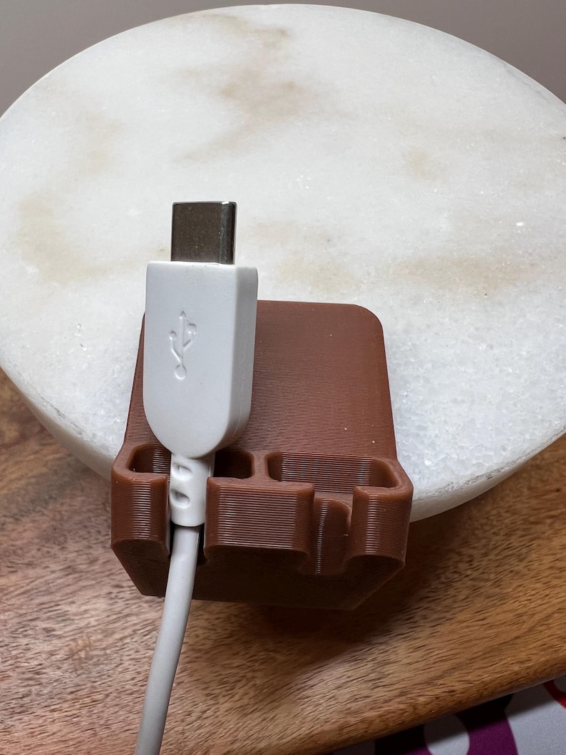 May include: A brown, 3D-printed cable organizer stand featuring three distinct slots for holding cords. A white USB-C cable is inserted into the leftmost slot, with the connector head facing upward, showcasing the standard USB symbol on its casing.