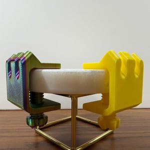 May include: Two C-clamps attached to a circular white stone disc supported by a gold-colored metal wire stand. One clamp features a metallic iridescent finish with green and purple hues, while the other clamp is a solid matte yellow plastic.