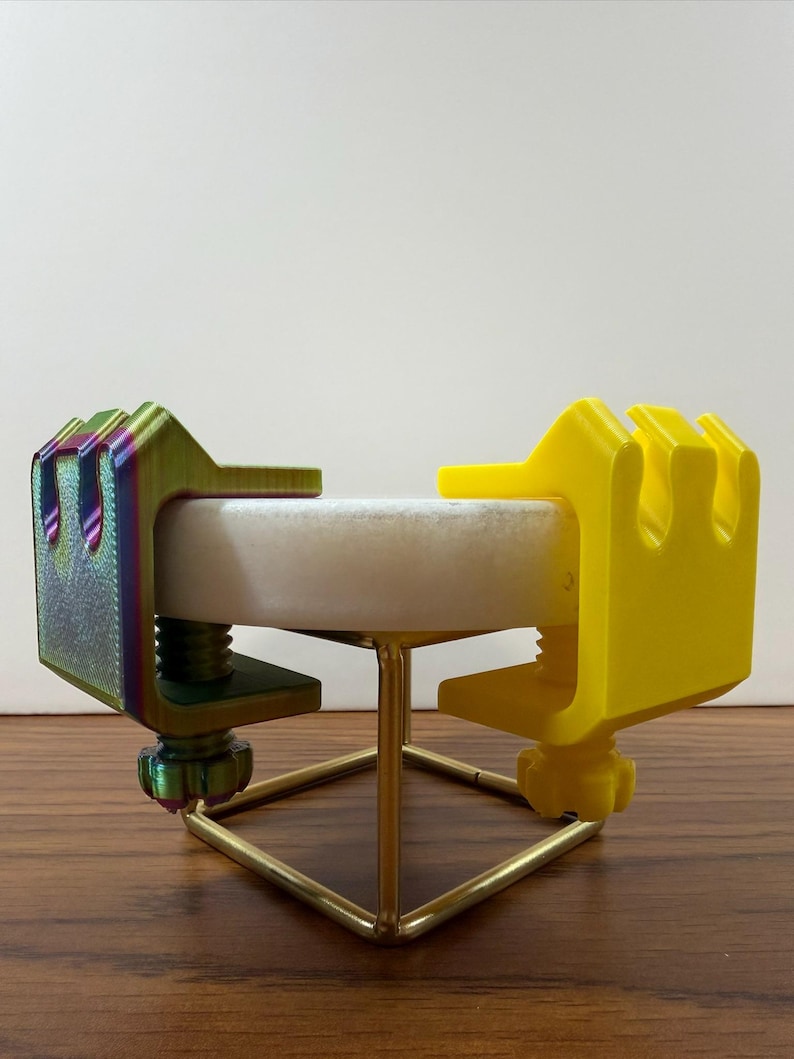 May include: Two C-clamps attached to a circular white stone disc supported by a gold-colored metal wire stand. One clamp features a metallic iridescent finish with green and purple hues, while the other clamp is a solid matte yellow plastic.