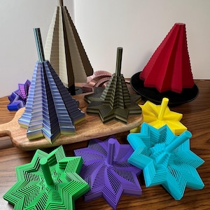 May include: A collection of colorful, geometric 3D-printed sculptures. The pieces include star-shaped and conical designs in various colors like green, purple, blue, yellow, and red. Some are on a wooden cutting board, others on a black plate.