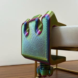 May include: A 3D-printed clamp featuring a vibrant iridescent finish that shifts between green, purple, and gold hues. The clamp has a U-shaped design with a threaded adjustment knob at the bottom, securely attached to a thin, gold-colored metal frame.