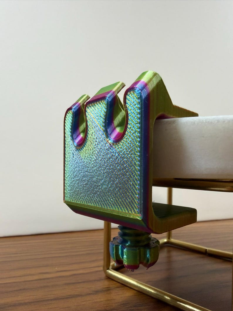 May include: A 3D-printed clamp featuring a vibrant iridescent finish that shifts between green, purple, and gold hues. The clamp has a U-shaped design with a threaded adjustment knob at the bottom, securely attached to a thin, gold-colored metal frame.