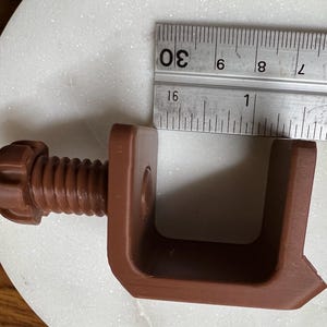 May include: A brown C-clamp with a threaded screw and a knob, positioned next to a metal ruler showing a measurement of approximately 1.25 inches for the inner opening width. The clamp has a U-shaped frame and a textured, matte finish.