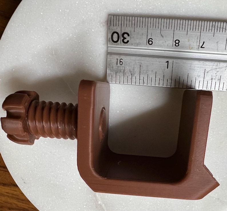 May include: A brown C-clamp with a threaded screw and a knob, positioned next to a metal ruler showing a measurement of approximately 1.25 inches for the inner opening width. The clamp has a U-shaped frame and a textured, matte finish.