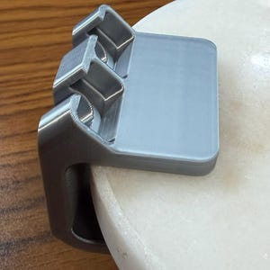 May include: A silver-colored, 3D-printed plastic clamp with a flat top surface and a vertical side piece. The object features three distinct, square-shaped recessed compartments along its top edge and is designed to grip onto a rounded edge.