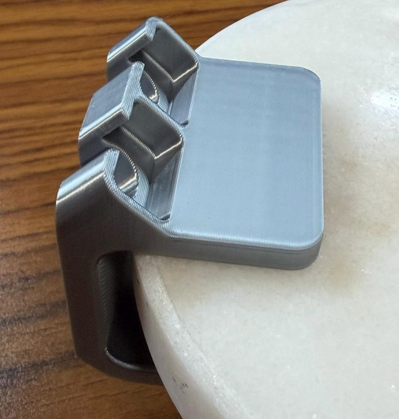 May include: A silver-colored, 3D-printed plastic clamp with a flat top surface and a vertical side piece. The object features three distinct, square-shaped recessed compartments along its top edge and is designed to grip onto a rounded edge.