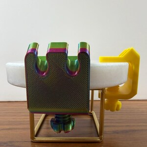 May include: A multi-colored, iridescent 3D-printed clamp with a three-pronged top and a threaded tightening knob, attached to a white circular disc. A separate bright yellow C-clamp is also attached to the side of the white disc.
