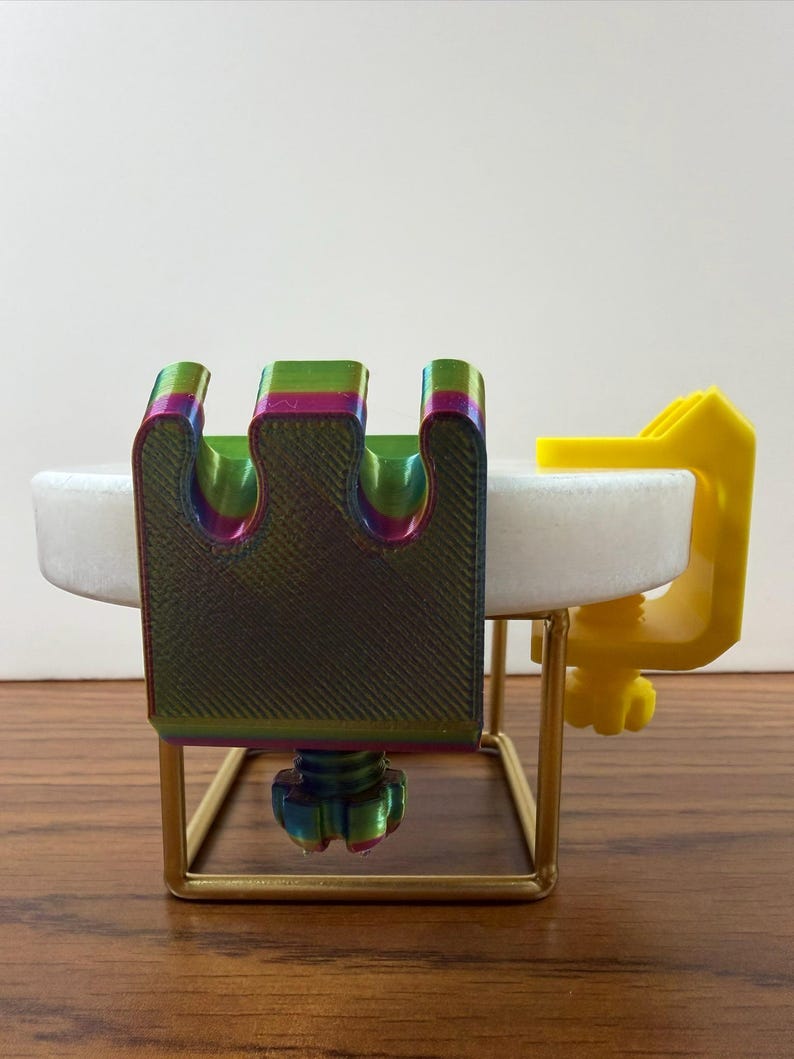 May include: A multi-colored, iridescent 3D-printed clamp with a three-pronged top and a threaded tightening knob, attached to a white circular disc. A separate bright yellow C-clamp is also attached to the side of the white disc.