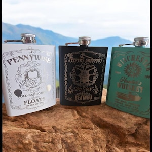 May include: Three stainless steel hip flasks with different designs. The first flask is white with a Pennywise design and the text "Old Fashioned Floats Derry, ME. USA". The second flask is black with a Jeepers Creepers design and the text "23rd Spring Beatngu". The third flask is teal with a Winchester design and the text "Since 1900 Hunter's Whiskey".