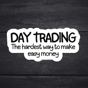 May include: White sticker with the text "DAY TRADING" in bold, black, uppercase letters. Below, in a smaller, handwritten-style font, it reads "The hardest way to make easy money." The sticker has a white, cloud-like border, set against a dark wood grain background.