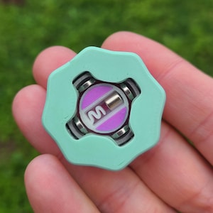 May include: A mint green fidget spinner with a purple center and the letter "M". The spinner has a unique hexagonal shape with rounded edges and metallic accents. The design is modern and eye-catching.