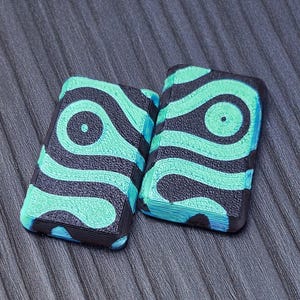 May include: Two rectangular objects with a black and teal wave pattern. The objects have rounded edges and a textured appearance. The teal design is a series of curved lines and circles. The background is a gray, ridged surface.