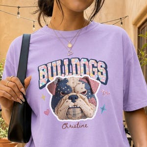 May include: A light purple short-sleeve t-shirt featuring a graphic of a bulldog with a star-spangled pattern on its head, the text BULLDOGS in a varsity font, and the name Christine written in cursive script below the dog illustration.