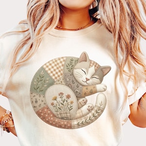 May include: A cream-colored short-sleeve t-shirt featuring a graphic of a curled-up cat. The cat illustration is composed of various patchwork-style segments in muted earth tones, including floral patterns, gingham checks, and solid beige.