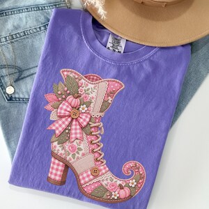 May include: A purple t-shirt featuring a graphic of a lace-up boot decorated with pink gingham patterns, floral motifs, and small pumpkins. The shirt is paired with light blue denim jeans, a tan wide-brimmed hat, and a delicate gold necklace.
