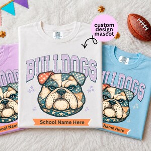 May include: Three t-shirts in light purple, white, and light blue, each featuring a graphic of a bulldog face with a star and plaid pattern. The text "BULLDOGS" appears above the bulldog, and "School Name Here" is printed on an orange banner below.