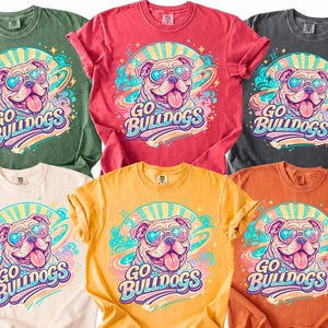 May include: Six short-sleeved t-shirts in olive green, red, charcoal gray, cream, yellow, and orange. Each shirt features a graphic of a bulldog wearing heart-shaped sunglasses with the text "GO BULLDOGS" in a colorful, swirling, retro-inspired design.