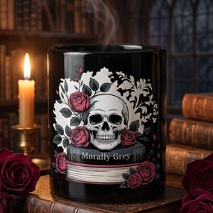 Op de afbeelding: Een zwarte keramische mok met een grafische illustratie van een menselijke schedel die rust op een boek met de titel Morally Grey, omringd door dieprode rozen en witte bloemaccenten. Er stijgt stoom op uit de mok, die een glanzende afwerking heeft.