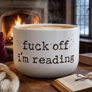 K&ouml;nnte beinhalten: Ein gro&szlig;er wei&szlig;er Keramikbecher gef&uuml;llt mit einem dampfenden Getr&auml;nk, mit dem schwarzen Aufdruck 'fuck off i'm reading' auf der Vorderseite. Der Becher hat eine glatte, abgerundete Form und steht auf einer Holzoberfl&auml;che mit Sprenkeln.