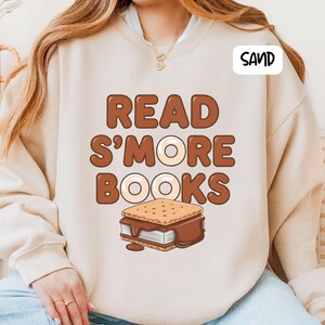 以下が含まれることがあります： 砂色のスウェットシャツ。茶色の文字で「READ S'MORE BOOKS」と書かれています。「MORE」の「O」はドーナツ型です。テキストの下にはスモアのグラフィックがあります。「SAND」という単語が右上隅の白いボックスにあります。