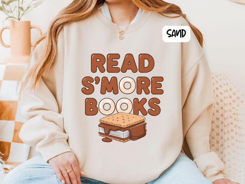 以下が含まれることがあります： 砂色のスウェットシャツ。茶色の文字で「READ S'MORE BOOKS」と書かれています。「MORE」の「O」はドーナツ型です。テキストの下にはスモアのグラフィックがあります。「SAND」という単語が右上隅の白いボックスにあります。