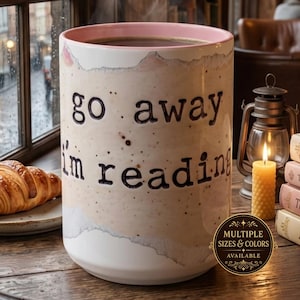 Könnte beinhalten: Ein zylindrischer Keramikbecher mit beiger, gesprenkelter Oberfläche und einem zartrosa Innenrand. Auf der Vorderseite ist der schwarze Text "go away i'm reading" aufgedruckt. Ein rundes goldenes Abzeichen zeigt "MULTIPLE SIZES & COLORS AVAILABLE".