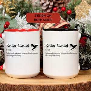 May include: Two white ceramic mugs with black text that reads "Rider Cadet" and a dragon graphic. One mug has a red handle and interior, the other has a black handle and interior. The mugs are on a wooden surface.