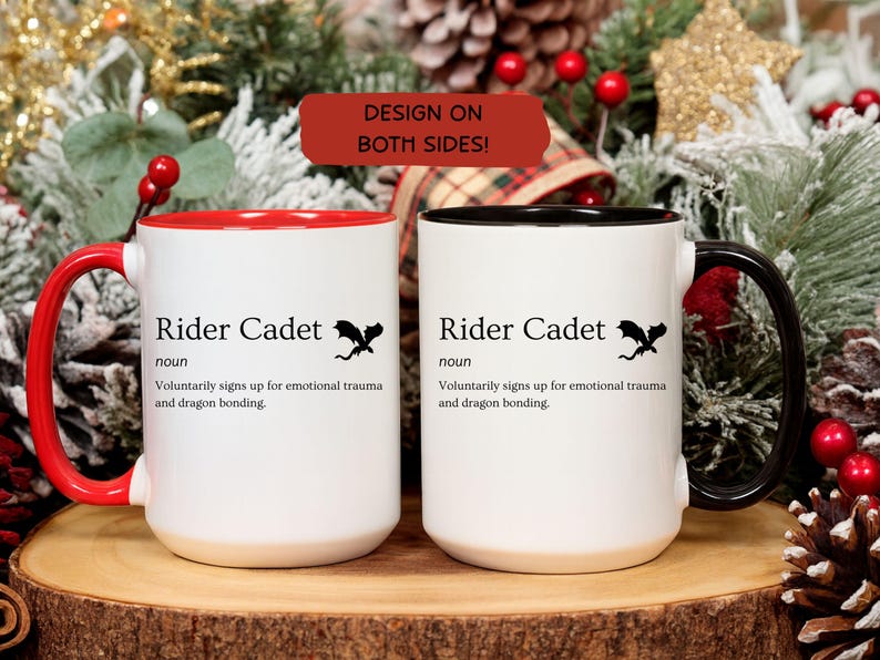 May include: Two white ceramic mugs with black text that reads "Rider Cadet" and a dragon graphic. One mug has a red handle and interior, the other has a black handle and interior. The mugs are on a wooden surface.