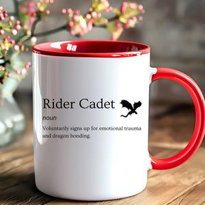 May include: White ceramic mug with a red interior and handle. The mug features the text "Rider Cadet" with a dragon silhouette. The definition below reads "Voluntarily signs up for emotional trauma and dragon bonding."