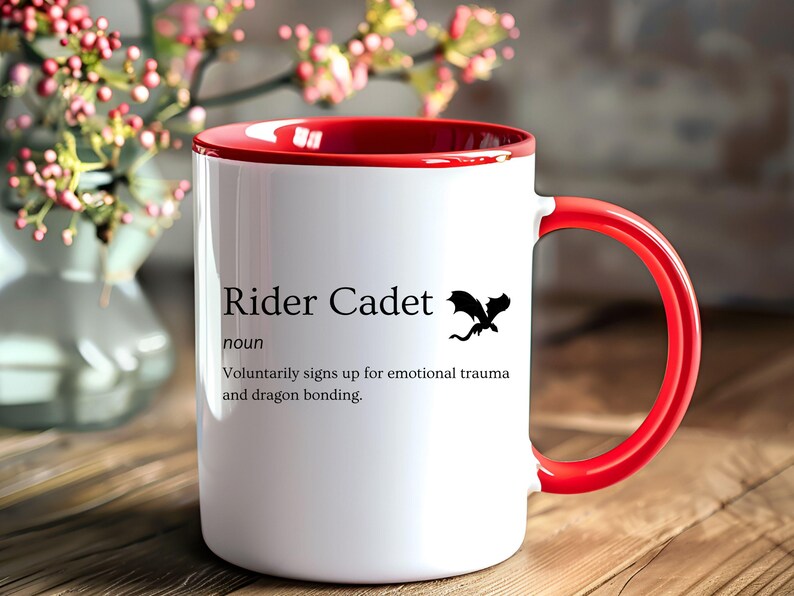 May include: White ceramic mug with a red interior and handle. The mug features the text "Rider Cadet" with a dragon silhouette. The definition below reads "Voluntarily signs up for emotional trauma and dragon bonding."