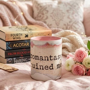 May include: A stack of books with titles including "Fourth Wing" and "ACOTAR" sits beside a ceramic mug with the text "romantasy ruined me." Pink and white roses and a wooden bookmark complete the cozy scene. The books and mug are on a pink blanket.