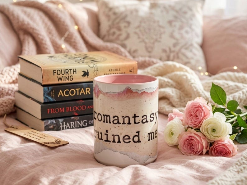 May include: A stack of books with titles including "Fourth Wing" and "ACOTAR" sits beside a ceramic mug with the text "romantasy ruined me." Pink and white roses and a wooden bookmark complete the cozy scene. The books and mug are on a pink blanket.