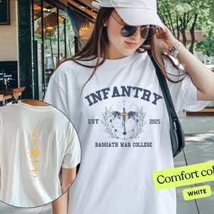 Puede incluir: Camiseta blanca con un estampado gr&aacute;fico azul de un drag&oacute;n con alas y el texto "INFANTRY EST 2025 BASSGIATH WAR COLLEGE". El texto "Comfort colors WHITE" es visible en la esquina inferior derecha de la imagen.