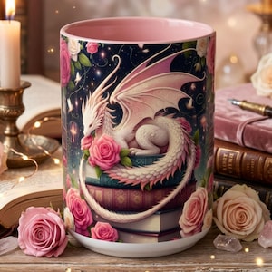 Może przedstawiać: Ceramiczny kubek z r&oacute;żowym wnętrzem i fantazyjnym wzorem smoka. Kubek przedstawia białego smoka z r&oacute;żowymi skrzydłami i r&oacute;żami. Tło jest ciemnoniebieskie z gwiazdami i kwiatowymi akcentami. Kubek jest otoczony książkami i r&oacute;żami.