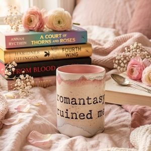 May include: A stack of books with titles like "A Court of Thorns and Roses" and "Fourth Wing" sits near a mug that reads "romantasy ruined me." The scene is decorated with pink flowers and a spoon, all on a soft, pink blanket.