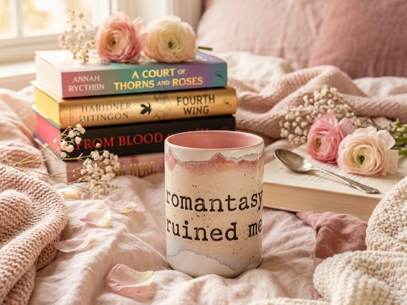 May include: A stack of books with titles like "A Court of Thorns and Roses" and "Fourth Wing" sits near a mug that reads "romantasy ruined me." The scene is decorated with pink flowers and a spoon, all on a soft, pink blanket.