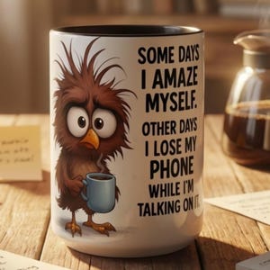 Könnte beinhalten: Ein weißer Keramikbecher mit schwarzem Innenrand, bedruckt mit einem braunen Cartoon-Vogel mit struppigem Gefieder, der eine kleine blaue Tasse hält. Der Text lautet: SOME DAYS I AMAZE MYSELF. OTHER DAYS I LOSE MY PHONE WHILE I'M TALKING ON IT.