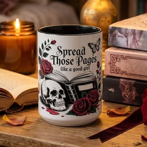 May include: White ceramic mug with a black interior, featuring a skull, roses, and the text "Spread Those Pages like a good girl." The mug is surrounded by books, a candle, and a red rose.