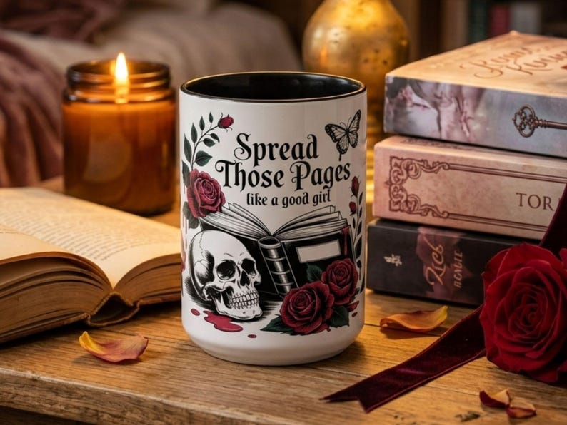 May include: White ceramic mug with a black interior, featuring a skull, roses, and the text "Spread Those Pages like a good girl." The mug is surrounded by books, a candle, and a red rose.