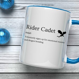 May include: White ceramic mug with a blue handle and rim, featuring the text "Rider Cadet" and a dragon silhouette. The mug's definition humorously describes the role. Decorative blue spheres are in the background.