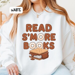 以下が含まれることがあります： 白いスウェットシャツ。茶色の文字で「READ S'MORE BOOKS」と書かれています。テキストの下にはスモアのグラフィックがあります。スウェットシャツはカジュアルなスタイルです。