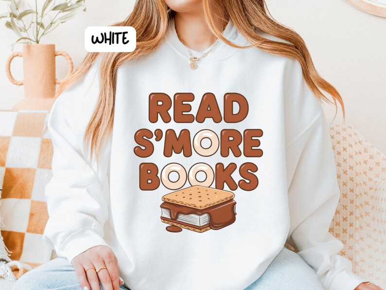 以下が含まれることがあります： 白いスウェットシャツ。茶色の文字で「READ S'MORE BOOKS」と書かれています。テキストの下にはスモアのグラフィックがあります。スウェットシャツはカジュアルなスタイルです。