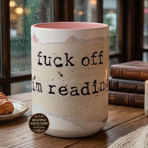 May include: A white ceramic mug with a pink interior featuring the black text "fuck off i'm reading" in a typewriter-style font. The exterior has a textured, paper-like appearance with torn edges. A small circular badge states "MULTIPLE SIZES & COLORS AVAILABLE".