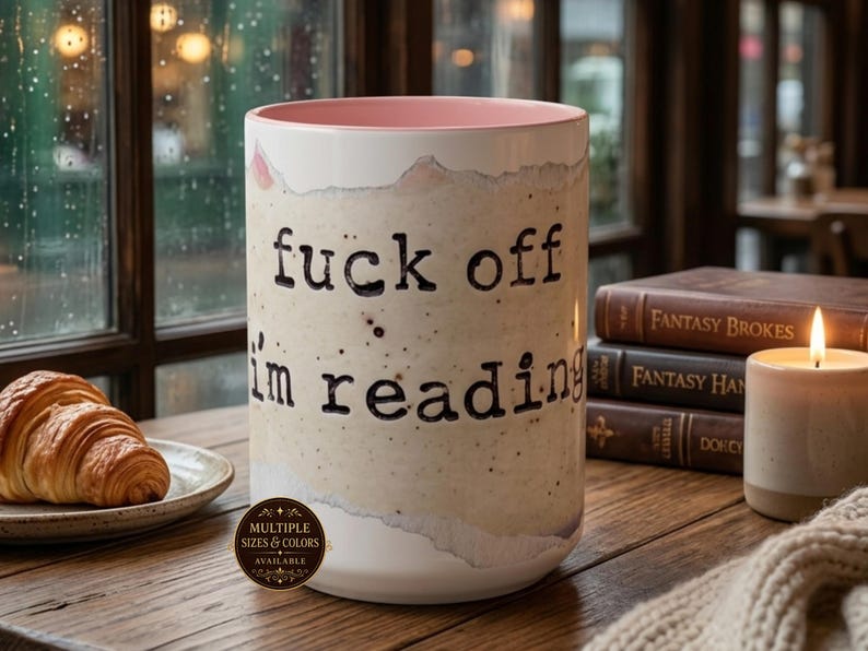 K&ouml;nnte beinhalten: Ein wei&szlig;er Keramikbecher mit rosa Innenseite und dem schwarzen Text "fuck off i'm reading" in Schreibmaschinenschrift. Die Au&szlig;enseite hat eine texturierte, papierartige Optik mit ausgefransten R&auml;ndern. Ein kleines rundes Abzeichen zeigt "MULTIPLE SIZES & COLORS AVAILABLE."